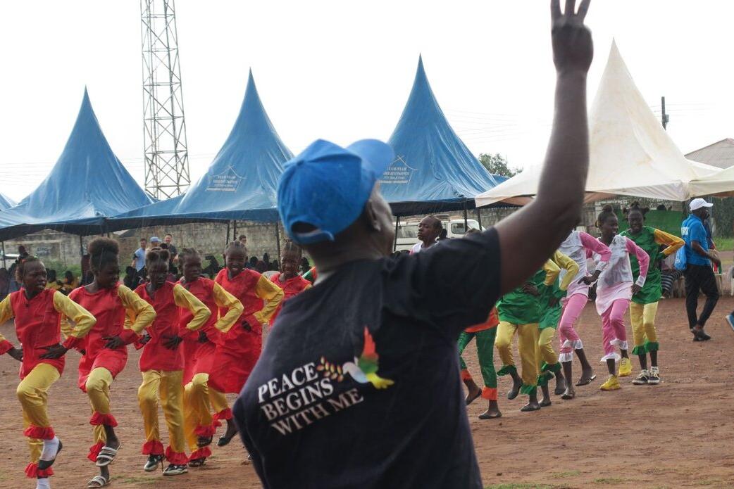 Wau’s third annual marathon, gathering over 2,000 participants, launched the UNMISS campaign "Peace Begins With Me” in Western Bahr El Ghazal. Outdoor event with a person wearing a shirt that reads “Peace Begins With Me” raising a hand, while groups of performers in colorful outfits dance in formation near blue tents.