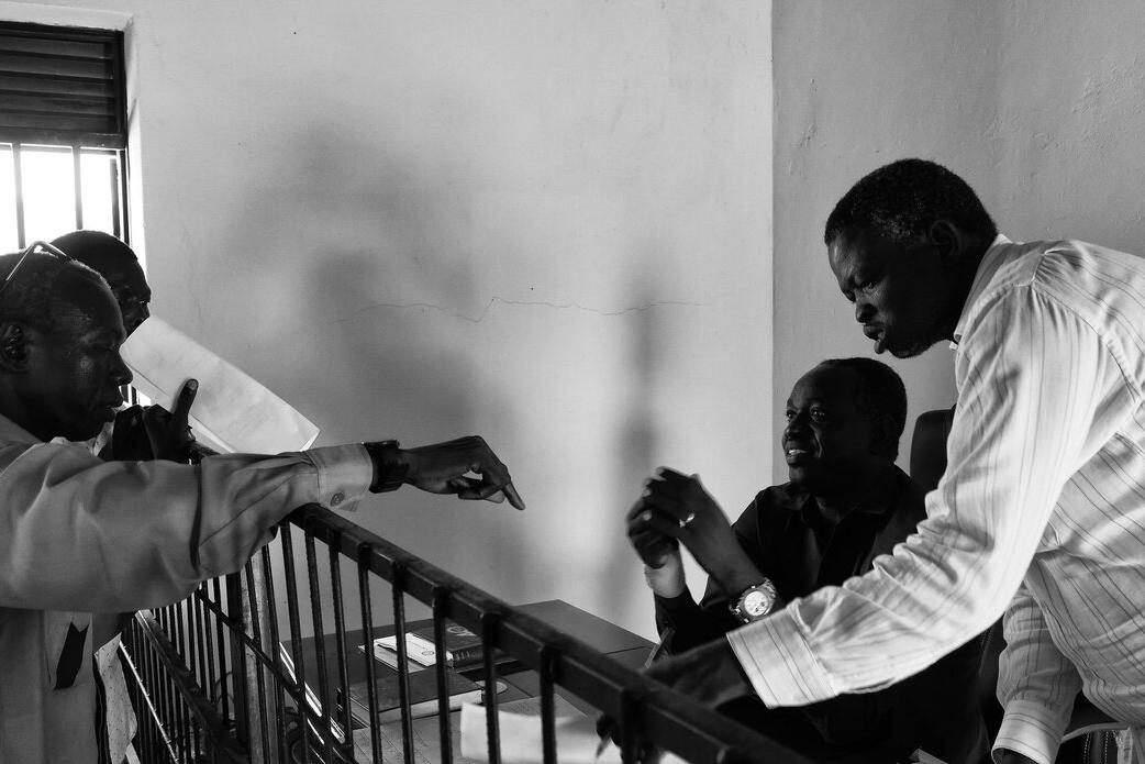 Three people engaged in discussion across a metal barrier inside a room with a barred window, papers and a notebook visible on the desk