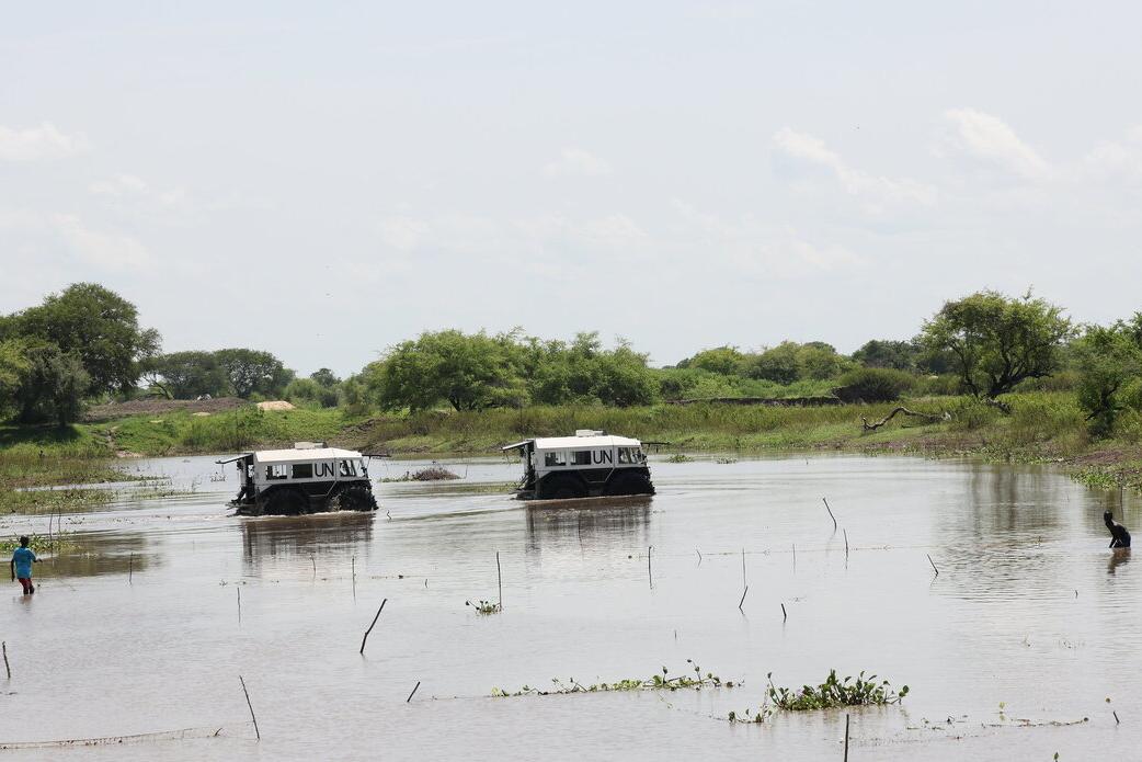 Two UN-marked armored vehicles driving through a shallow water body, with trees and vegetation in the background and two people standing in the water nearby.