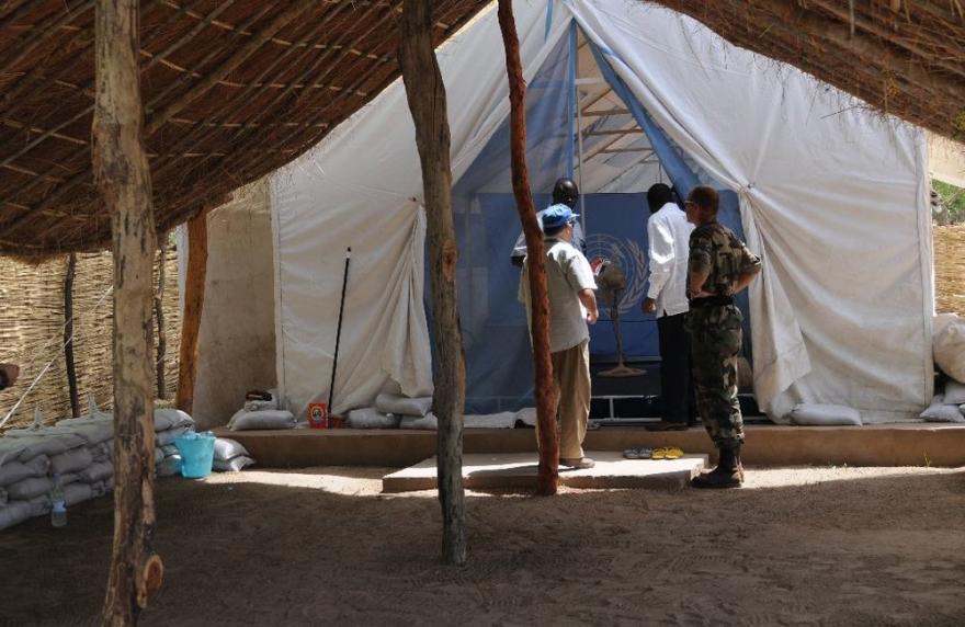 Three people stand inside a large white tent with a UN emblem, under a thatched roof, surrounded by sandbags and basic supplies.
